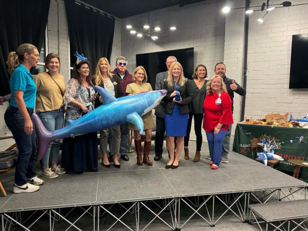 Nassau Education Foundation supporters and educators pose onstage with a large blue shark prop during a Shark Grant award presentation.