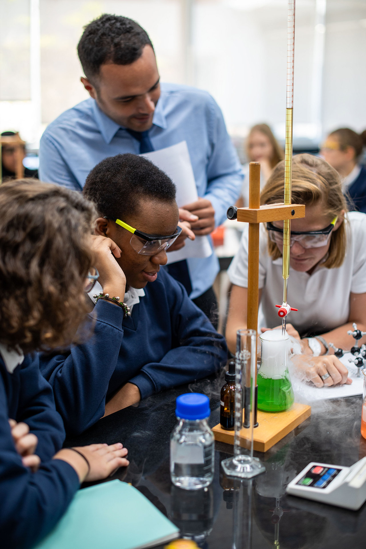 Teacher observes students wearing safety goggles as they conduct a hands-on chemistry experiment with laboratory equipment in the classroom.

Description: Students work together on a classroom science experiment.