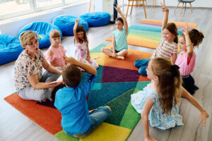 Teacher seated with young students on a colorful classroom rug as several children raise their hands during a group lesson.