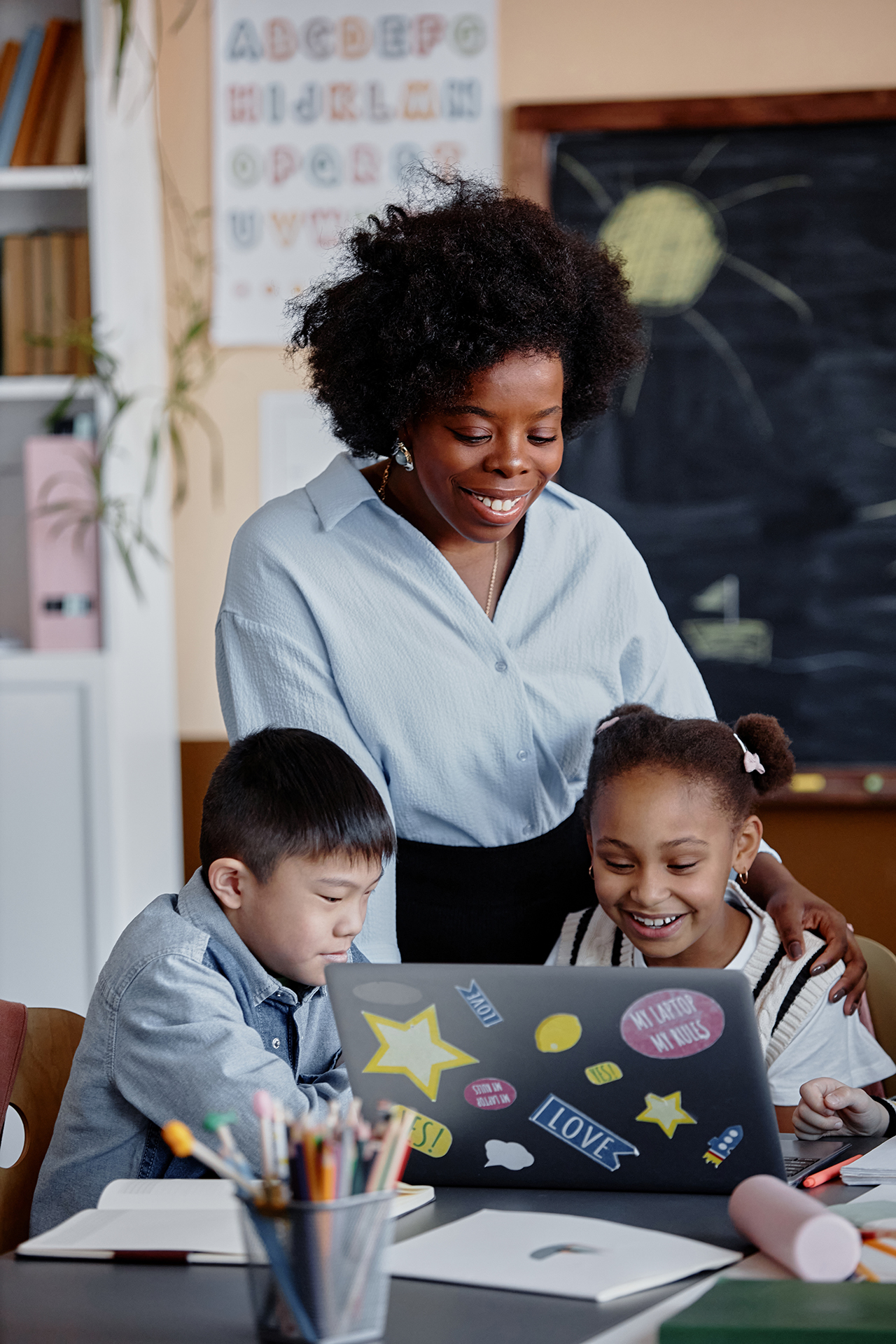 Teacher smiles while helping two elementary students work together on a laptop in the classroom.